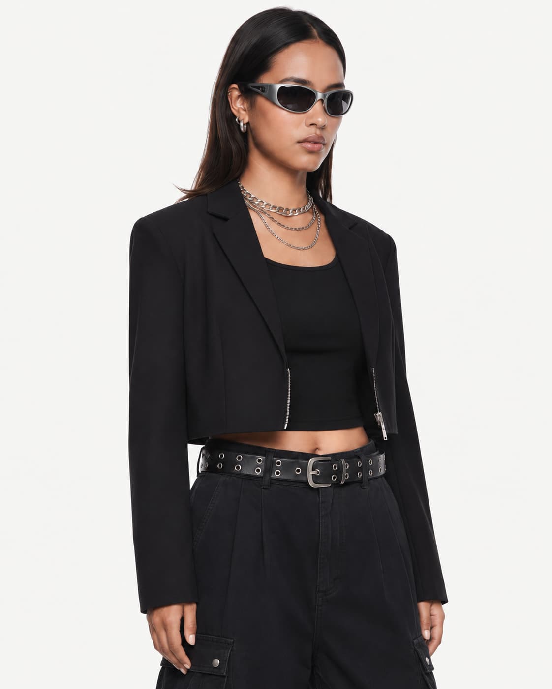 Close-up — the blazer's zipped cropped hem, layered chains and studded belt at waist height.