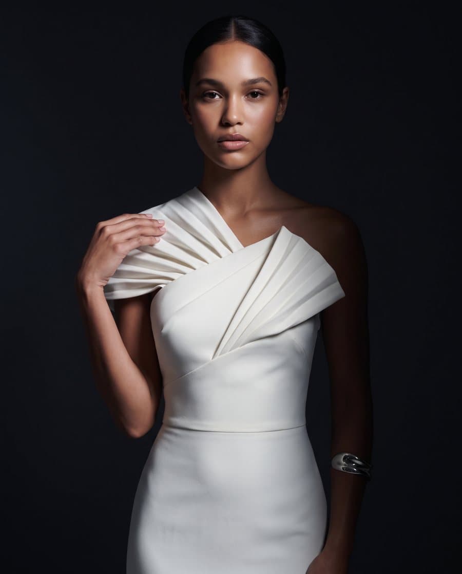 Ivory pleated one-shoulder gown lit by a single narrow spotlight on near-black