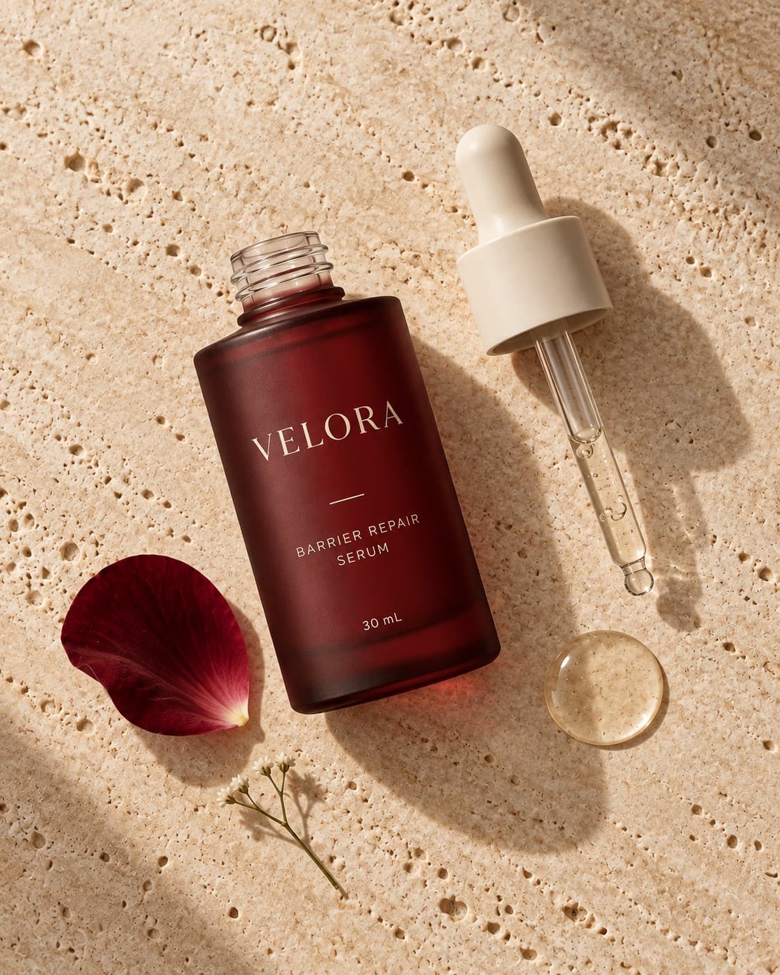 Overhead flat-lay — the open serum bottle, its pipette, a rose petal and a bead of serum on travertine.