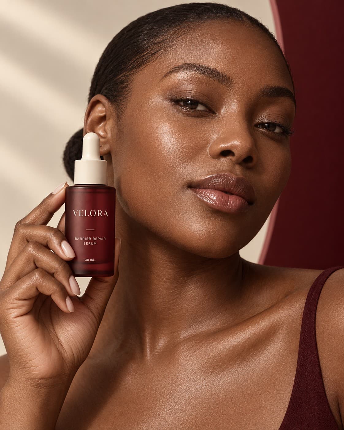 Beauty portrait — a model holding the VELORA serum beside her face in warm directional light.