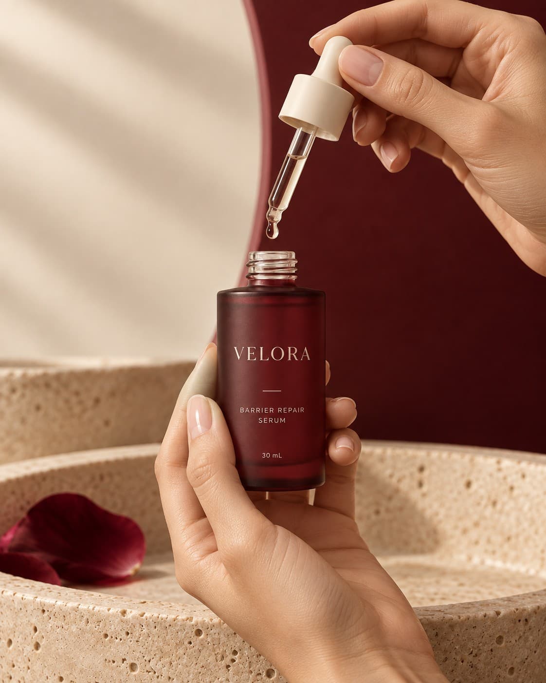 Two hands opening the VELORA serum over a travertine basin, a drop suspended at the pipette tip.