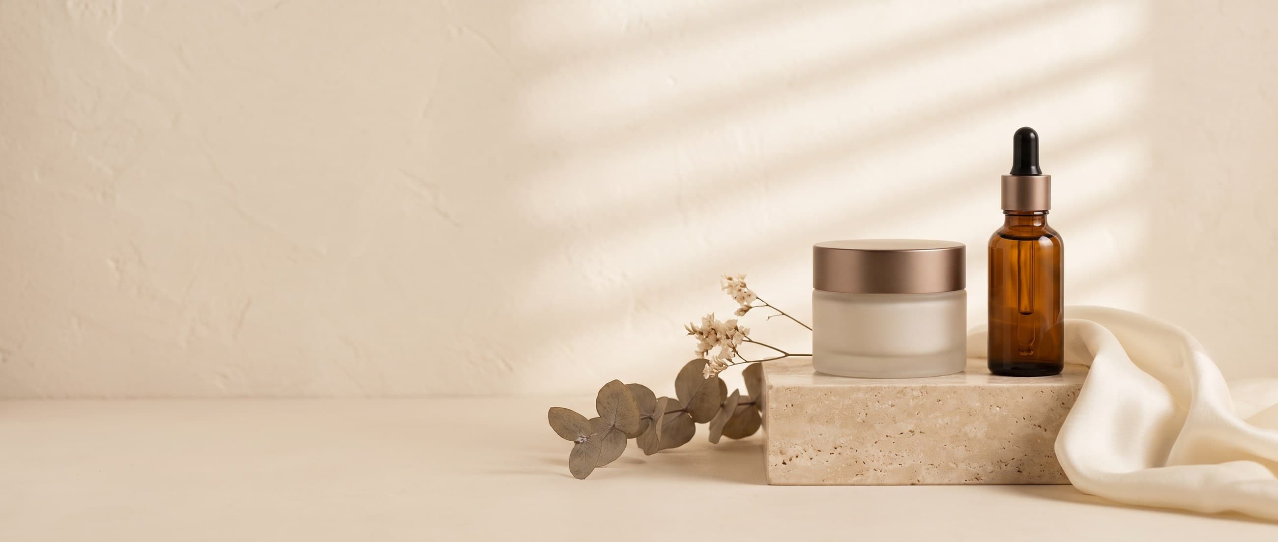 Beauty campaign — unlabeled skincare crème and serum on a travertine pedestal in soft daylight